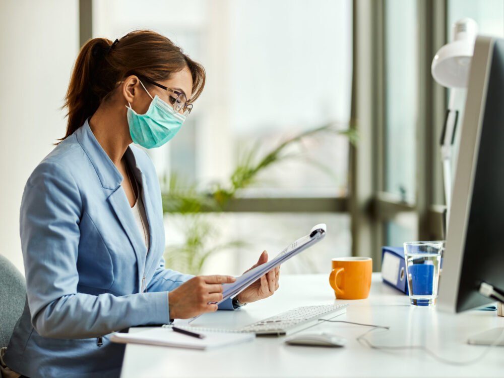 Female entrepreneur with face mask analyzing business reports in Young businesswoman wearing protective face mask while going through reports and working in the office.