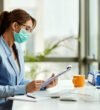 Young businesswoman wearing protective face mask while going through reports and working in the office.