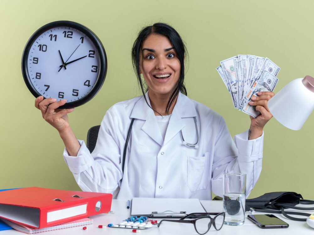 excited young female doctor wearing medical robe with stethoscope sits at desk with medical tools holding wall clock with cash isolated on olive green background excited young female doctor wearing medical robe with stethoscope sits at desk with medical tools holding wall clock with cash isolated on olive green background